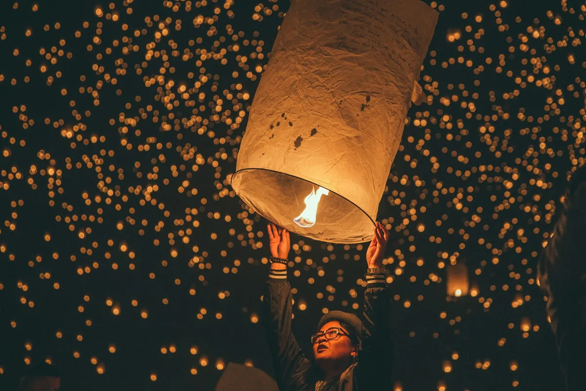 A traveler releasing a glowing paper sky lantern into a night sky full of floating lanterns at Chiang Mai's Yi Peng festival